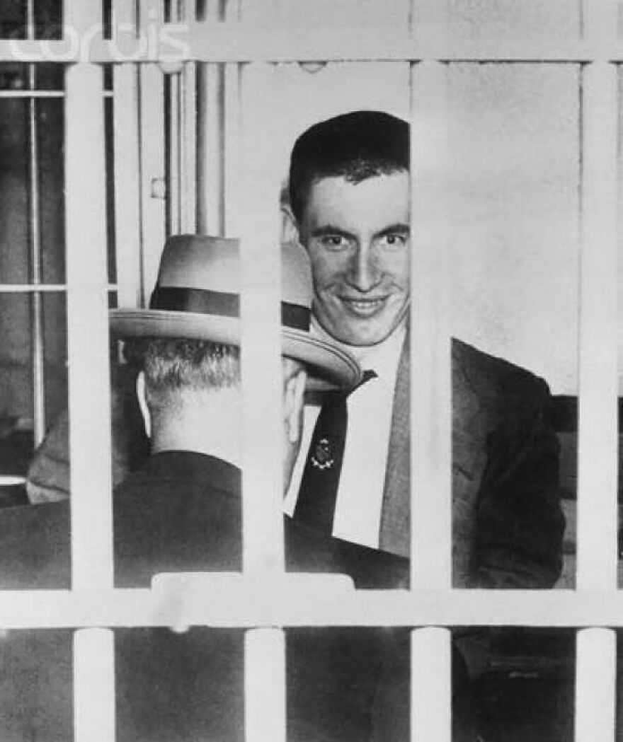 "Historical photo of a man smiling behind bars while talking to another man in a hat."