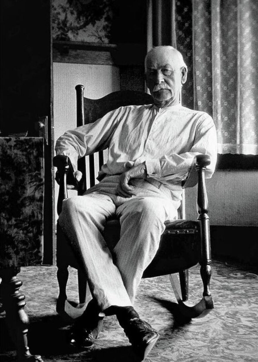 Elderly man in a rocking chair, wearing a light shirt and pants, in a historical photo setting.