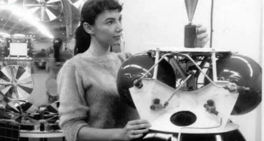 Woman in a historical photo examining a vintage machine, wearing a sweater, with mechanical parts visible.