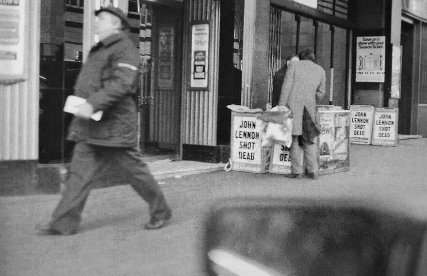 Black and white historical photo showing a street scene with newspapers displaying "John Lennon Shot Dead" headline.