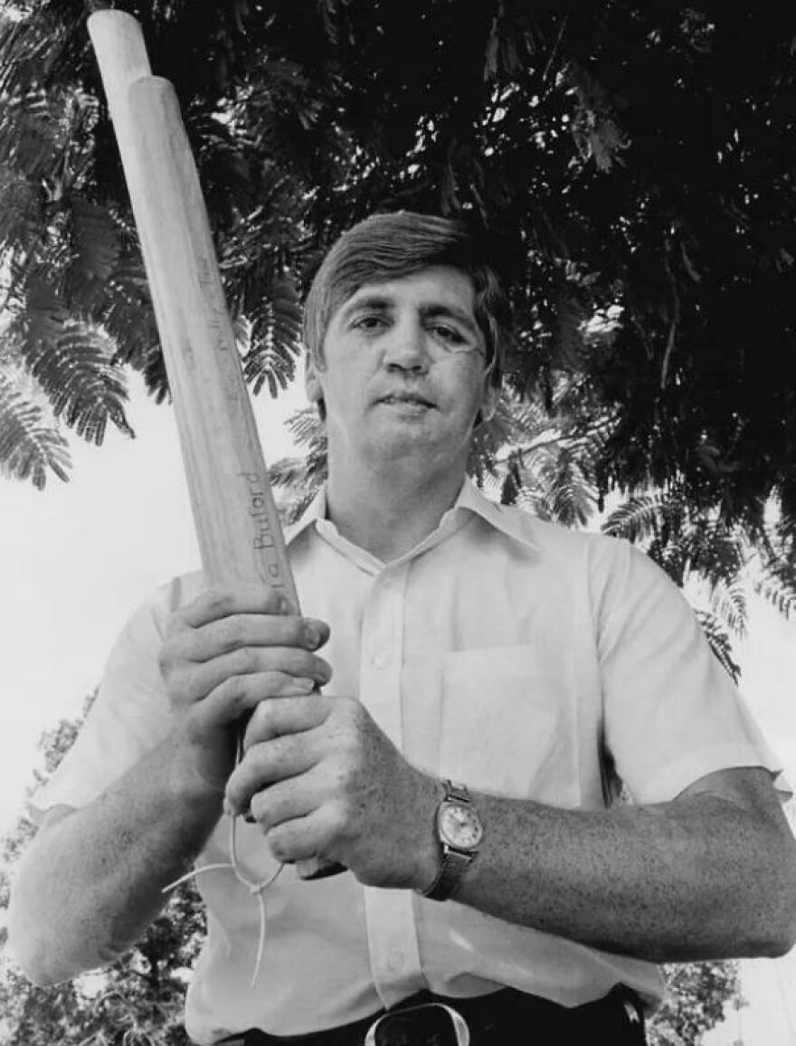 Historical photo of a man holding a large wooden club, wearing a watch and short-sleeved shirt, standing under a tree.