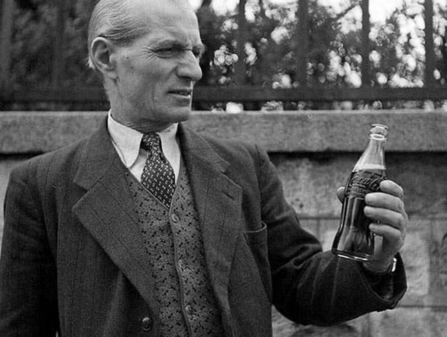 Man in a suit examining a Coca-Cola bottle outdoors, historical photo.