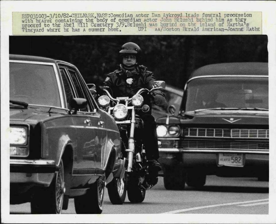 Motorcycle officer leads historical funeral procession with cars, 1982.