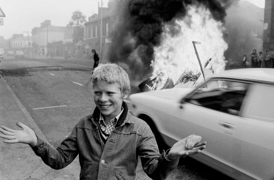 Boy smiling in front of a burning car during a historical protest, holding a stone amid the chaos.