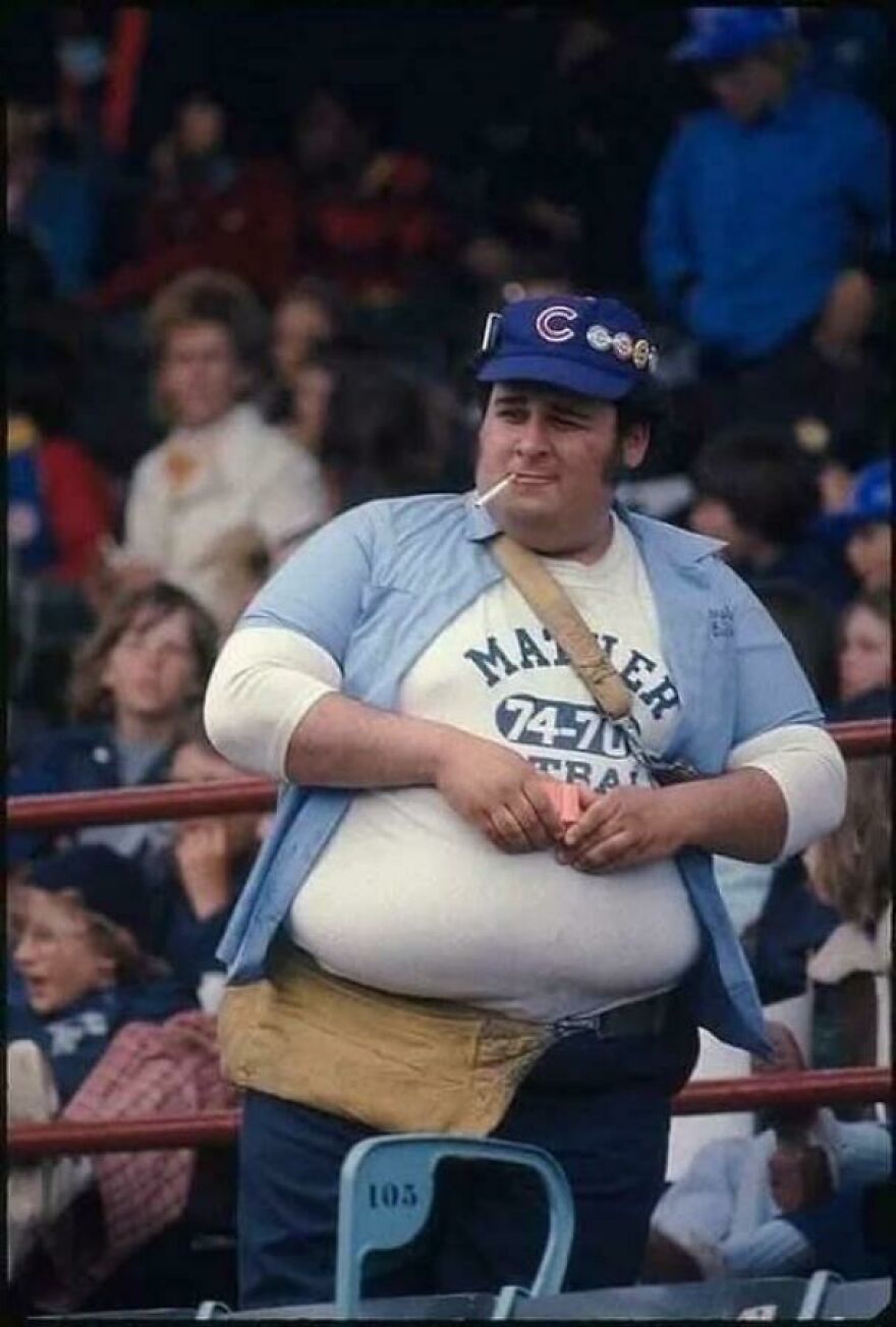 Stadium vendor wearing a cap and blue shirt, surrounded by a crowd, in a historical photo.