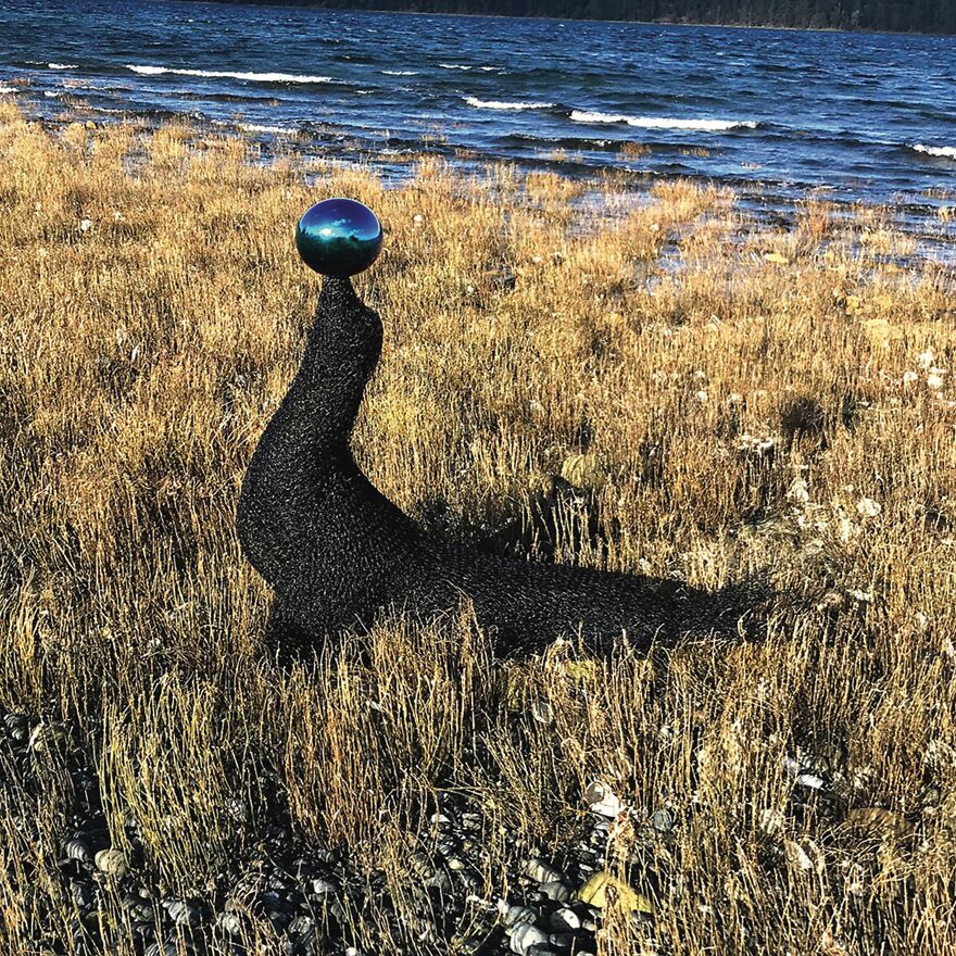 Chicken wire sculpture resembling a seal balancing a sphere by a lake.