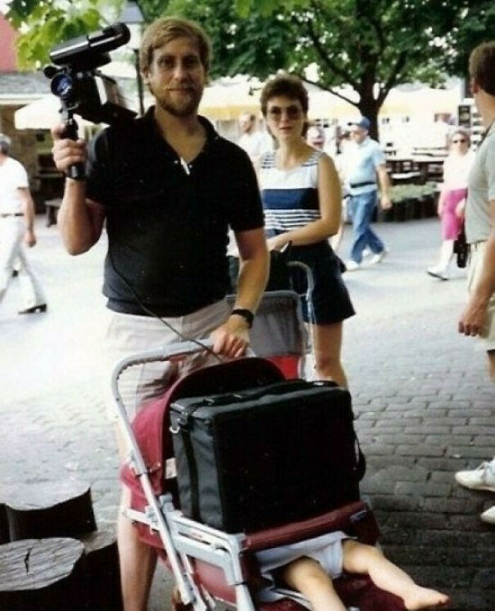 Man holding video camera, pushing stroller with a child’s legs sticking out, in an awkward family photo outdoors.