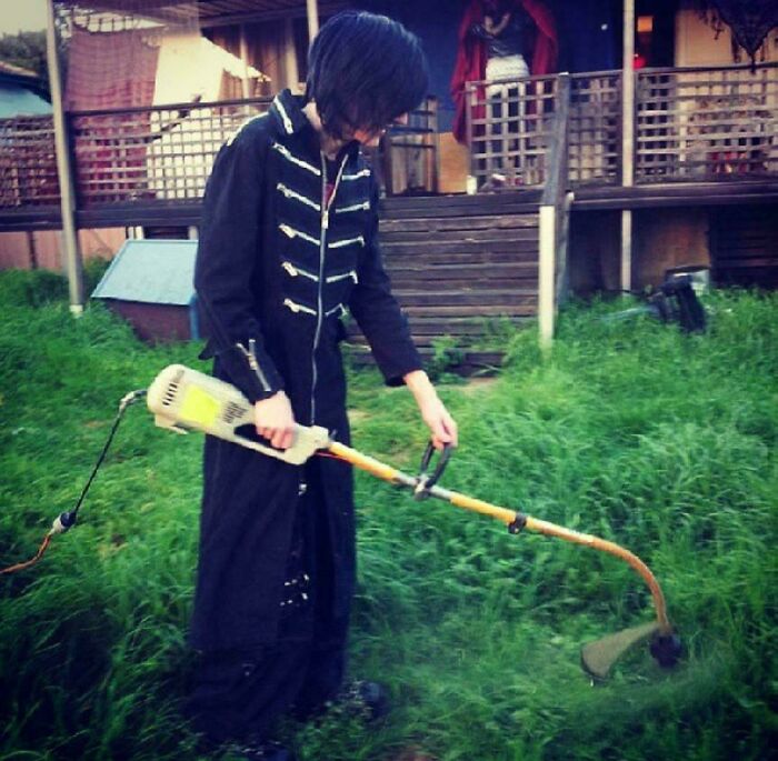 Person in a long black coat using a trimmer to cut overgrown grass in a backyard, a regrettable past moment captured.