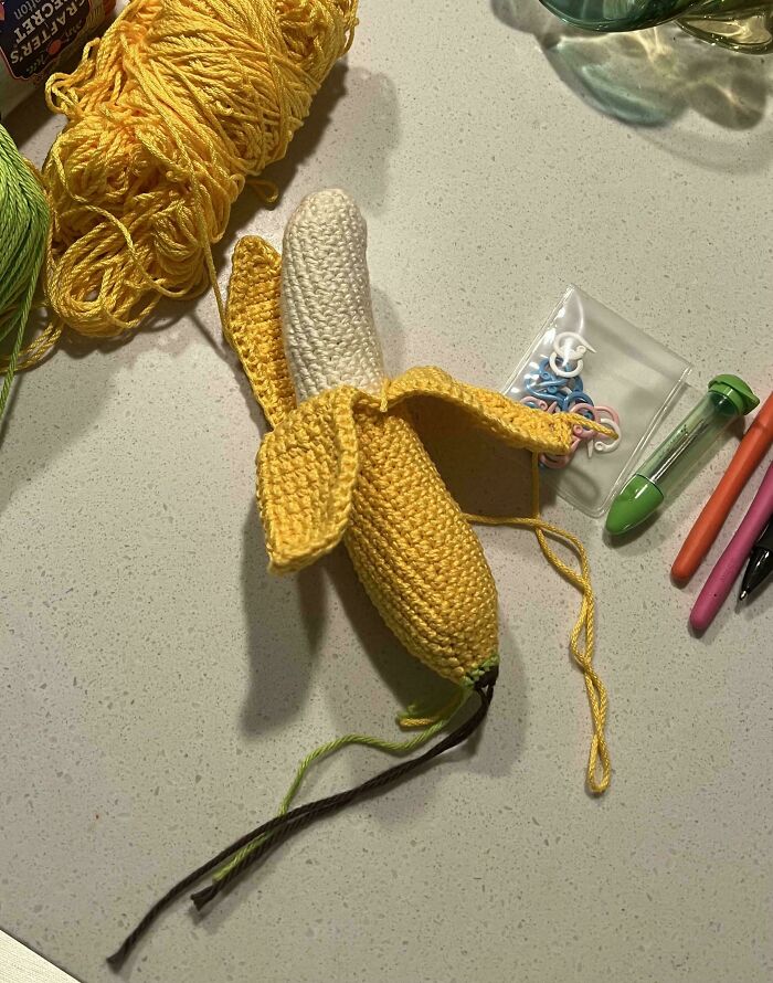 Crochet banana masterpiece in progress surrounded by yarn, crochet hooks, and stitch markers on a light surface.