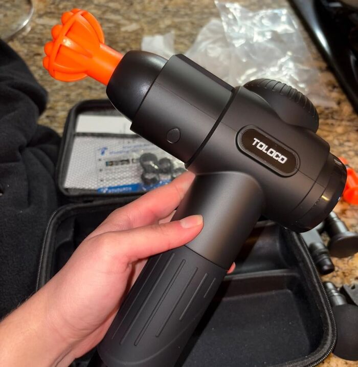 Hand holding a massage gun, ideal V-Day gift for long term couples, with an orange attachment and a carrying case nearby.