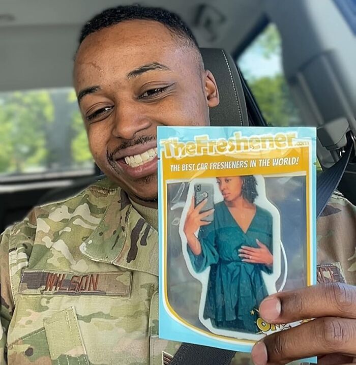 Man in military uniform holding a car freshener with a woman's photo, smiling inside a vehicle.