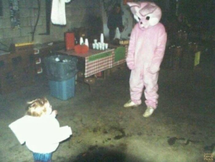 Child looking at adult in a pink bunny costume in a dimly lit room, an awkward family photo with a sweet vibe.