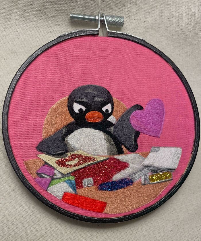 Embroidered penguin holding a purple heart with various colorful craft materials on a pink fabric background in embroidery hoop.