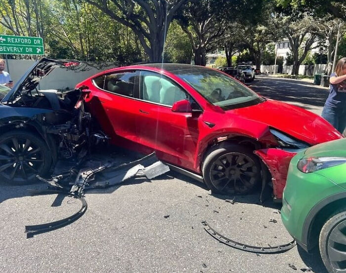 Three crashed cars, red, black, and green, with debris on the street from the accident. Bystanders look on.