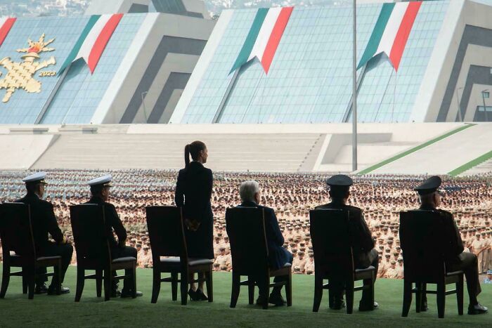 A large modern building with bold geometric patterns, viewed from a platform where officials observe a massive uniformed crowd.