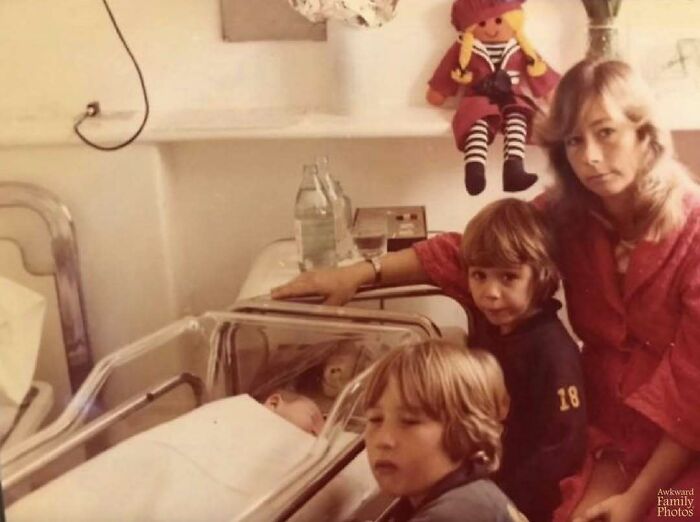 A family with two children and a mother posing awkwardly around a newborn baby in a clear hospital bassinet.
