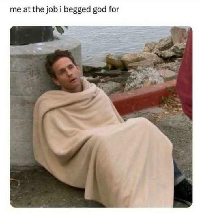 Person wrapped in a blanket, sitting on the ground, looking tired and funny near a waterfront.