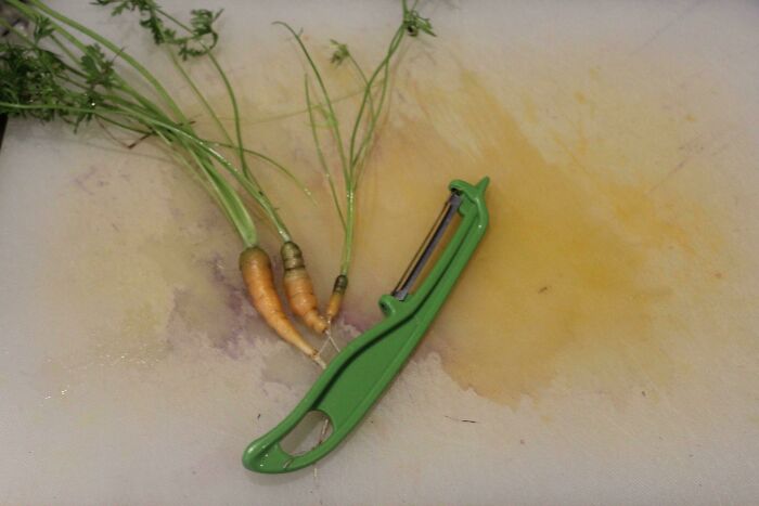 Three tiny carrots with green tops next to a green vegetable peeler on a cutting board, illustrating harvesting size humor.