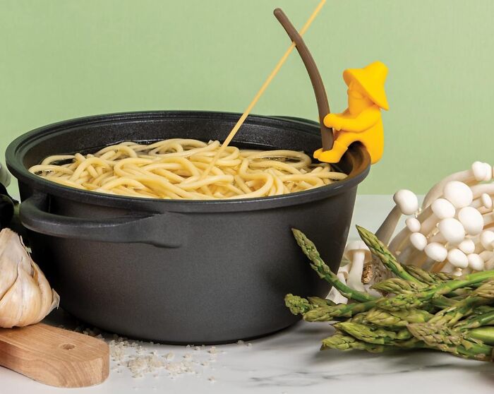 Weird kitchen item: a yellow figure fishing for noodles in a pot, surrounded by garlic, asparagus, and mushrooms.