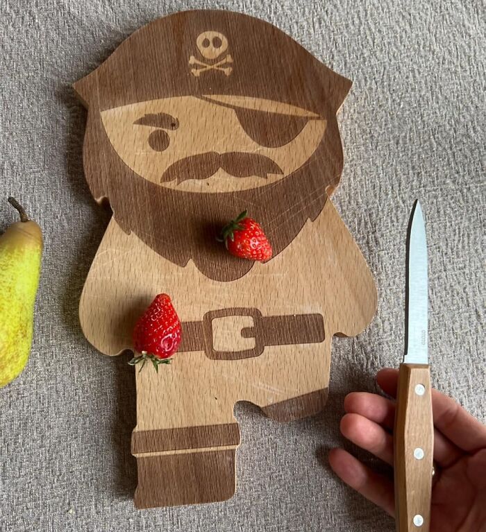 Pirate-shaped wooden cutting board with strawberries, alongside a pear and a paring knife; a unique kitchen item.