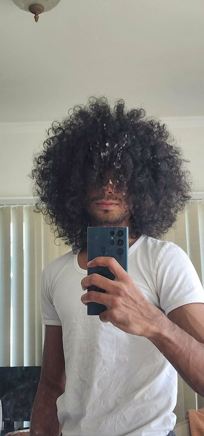 Man with fierceflow curly hair taking a mirror selfie wearing a white t-shirt in a bright room.