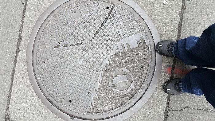 Manhole cover with city map design, showcasing cities-genius-solutions, sidewalk and feet in the frame.
