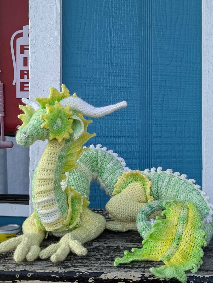 Crochet masterpiece of a detailed green and yellow dragon with white horns displayed on a wooden surface against a blue wall.