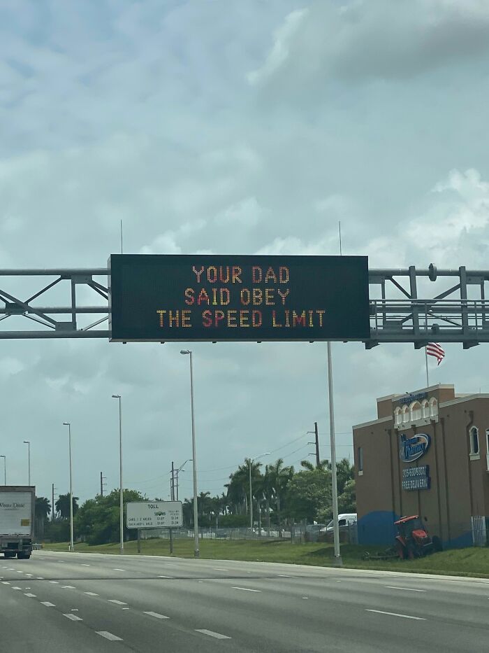Funny sign on highway reads "Your Dad Said Obey the Speed Limit" amidst traffic and buildings.