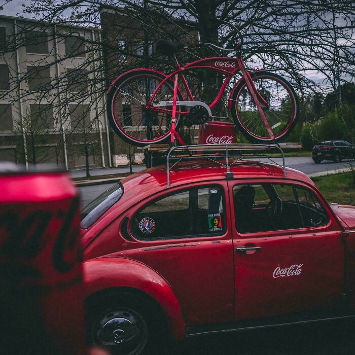 Red vintage car with Coca-Cola branding, featuring a matching bicycle on the roof, parked outdoors.