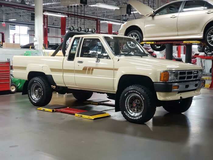 Vintage car in a garage, cream-colored and lifted for maintenance, surrounded by automotive tools and equipment.