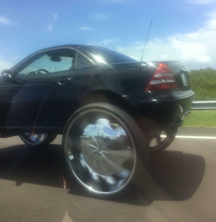 A black car with oversized wheels on a Florida highway, capturing a wild moment in the sunny weather.
