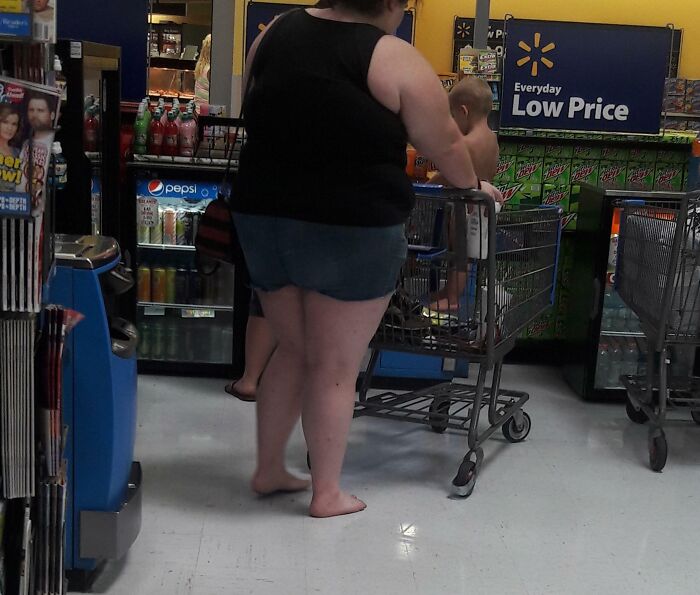 Barefoot shopper with child in cart in a Florida store aisle, surrounded by soft drink displays.