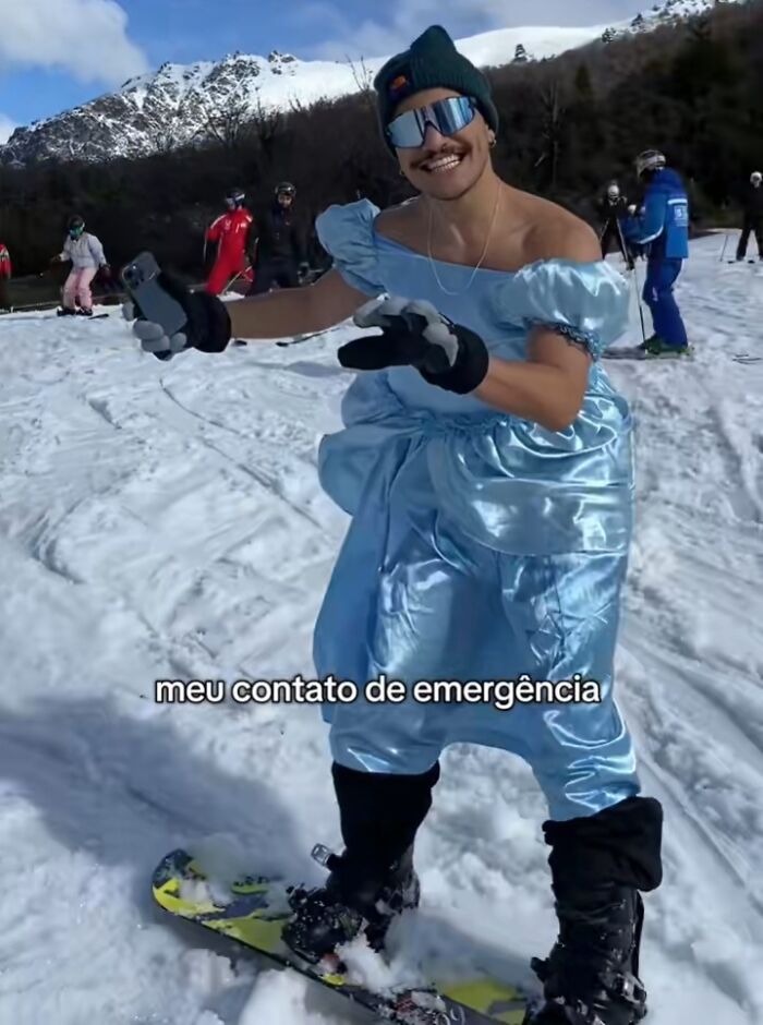 Man in a blue dress snowboarding on a snowy hill with mountains in the background, posing humorously for a photo.