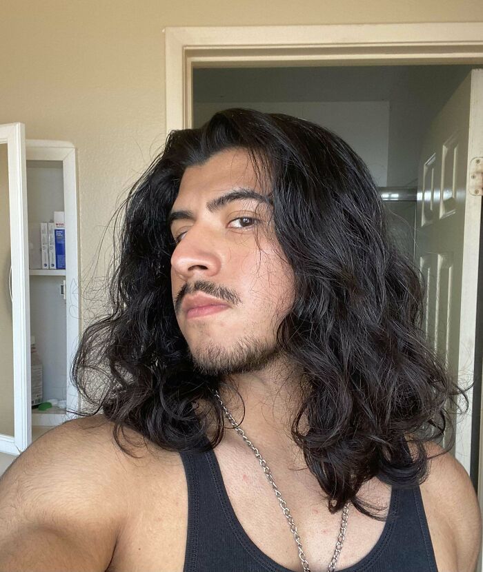 Man with fierceflow beautiful hair posing indoors wearing a black tank top and silver chain necklace.