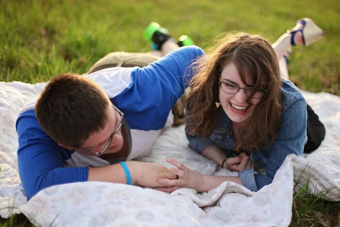 Couple sharing eye-opening moments, laughing while lying on a picnic blanket in a grassy field.