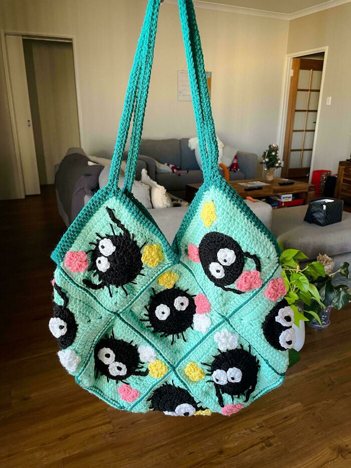 Crochet bag with black cartoon spider motifs and colorful flower accents, hanging in a cozy living room setting.