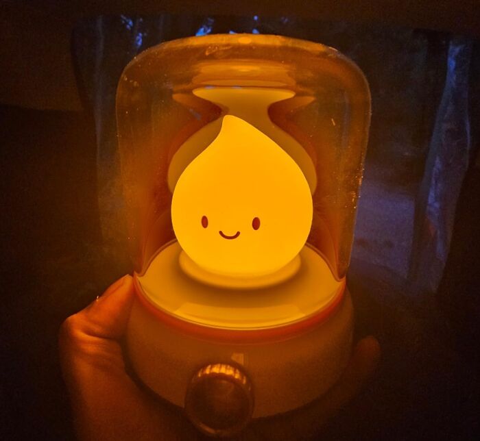 Hand holding a glowing, smiling, teardrop-shaped lamp under a glass cover, showcasing a weird, unique design.