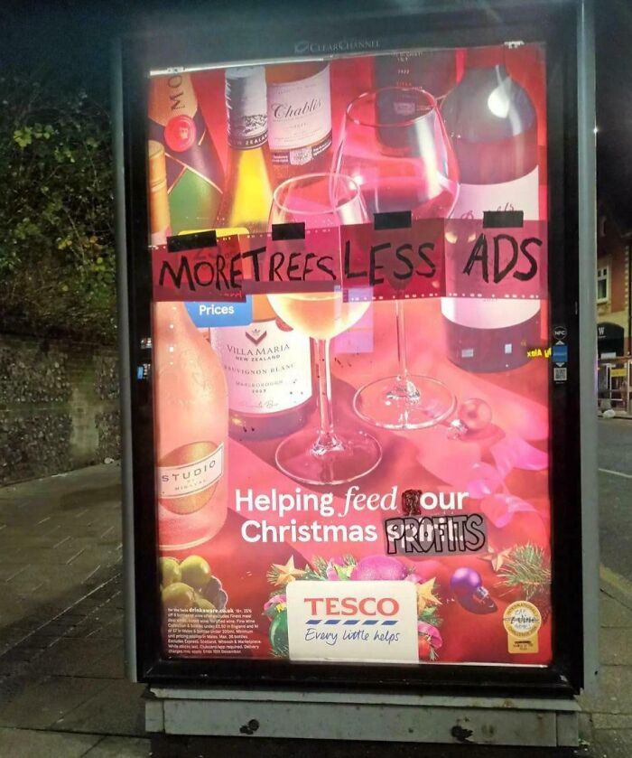 Bus stop ad altered with "MORE TREES LESS ADS" and "Helping feed our profits," reflecting anti-consumerism sentiment.