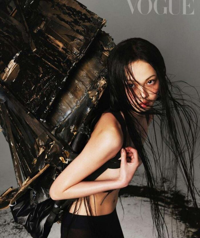 Person with long hair and dark clothing, carrying a large abstract object, symbolizing a rejection of consumerism.