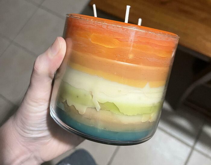 A multicolored candle in a clear jar, symbolizing consumerism, held in a hand against a tiled floor background.