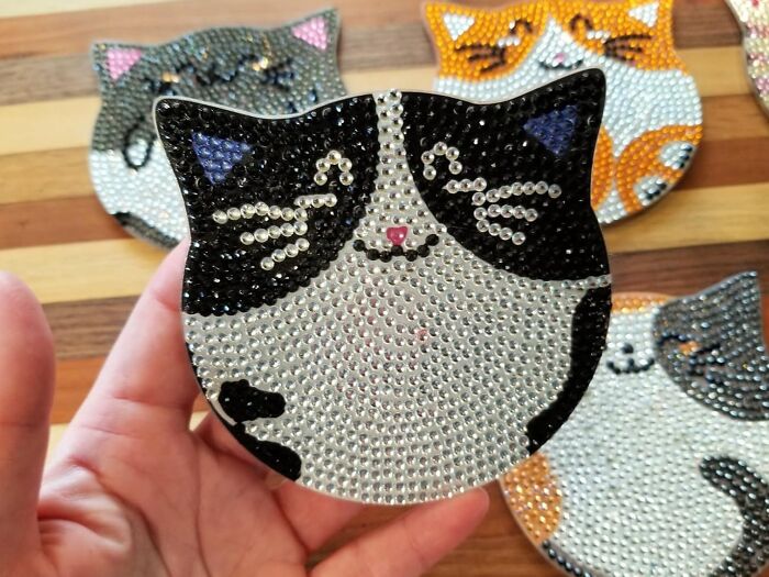 Hand holding a sparkling artistic cat face design, surrounded by other colorful cat coasters in the background.