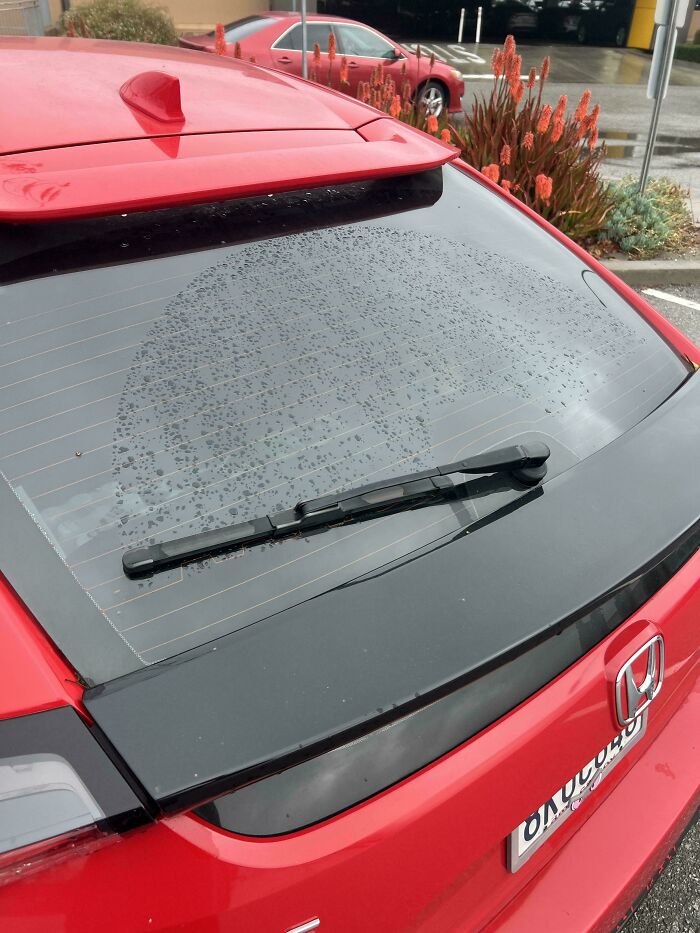 Red car rear windshield with water droplets cleared only in the wiper path, showing a moderately interesting pattern.