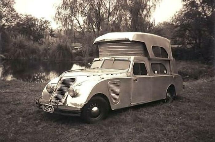 A unique, vintage car with a custom camper extension parked beside a tranquil lake.