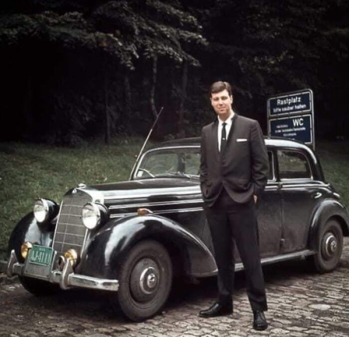 Man in a suit standing beside a classic vintage car with a forest background.