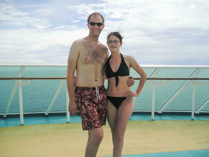 A couple wearing swimwear posing on a boat deck with the ocean and cloudy sky in the background, showing a regrettable past moment.
