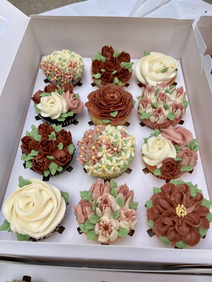 Box of twelve cupcakes decorated with incredible bakes featuring intricate floral designs in soft pink, white, and brown icing.