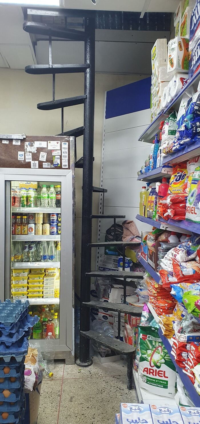 Narrow spiral death stairs in cluttered store aisle, surrounded by shelves and fridge.