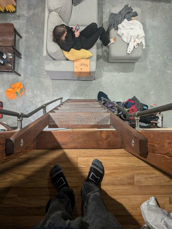 View from top of steep wooden "death stairs" with person sitting below on a couch, using a phone.
