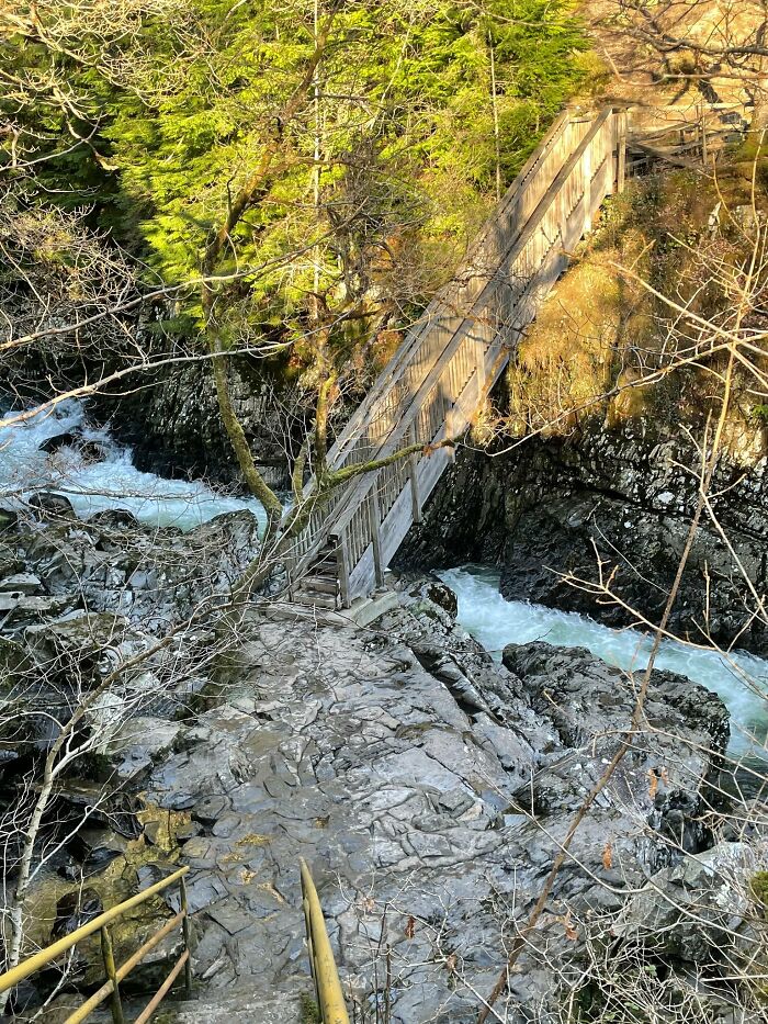Steep stairs leading to a narrow wooden bridge over a rocky river, surrounded by dense forest. Perfect "death stairs" example.