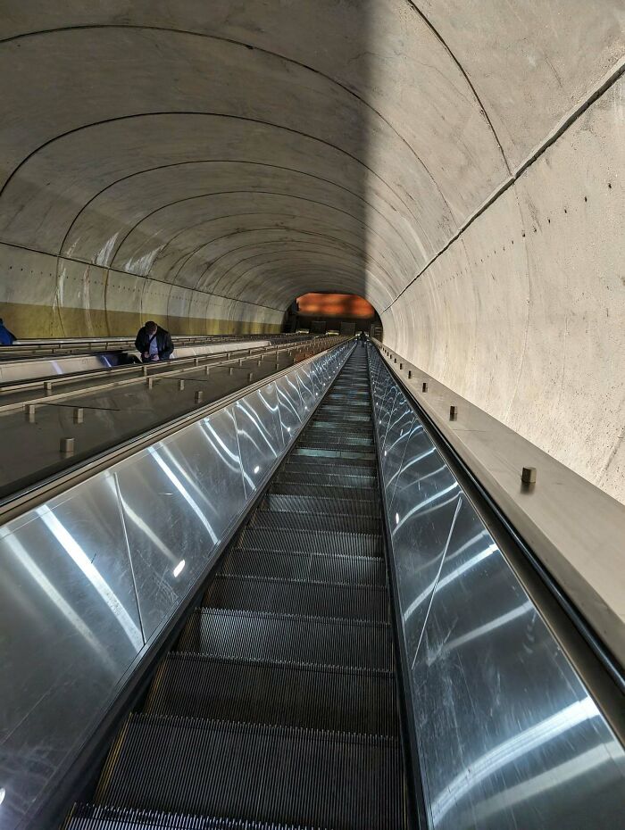Tall, steep escalator in a tunnel resembling 'death stairs,' leading down into a dimly lit underground area.